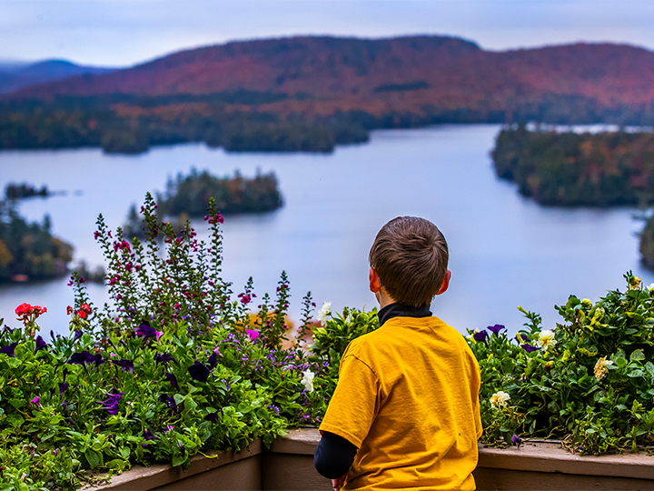 Un garçon regarde la vue sur le lac Blue Mountain et le feuillage changeant depuis la terrasse du Lake View Cafe. Visitez la région des Adirondacks dès aujourd'hui !