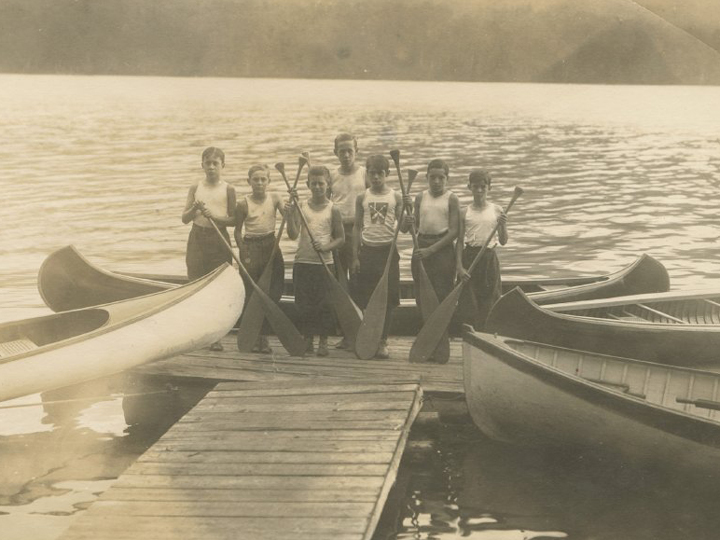 A vintage group of boys on the end of a dock holding paddles and surrounded by canoes.