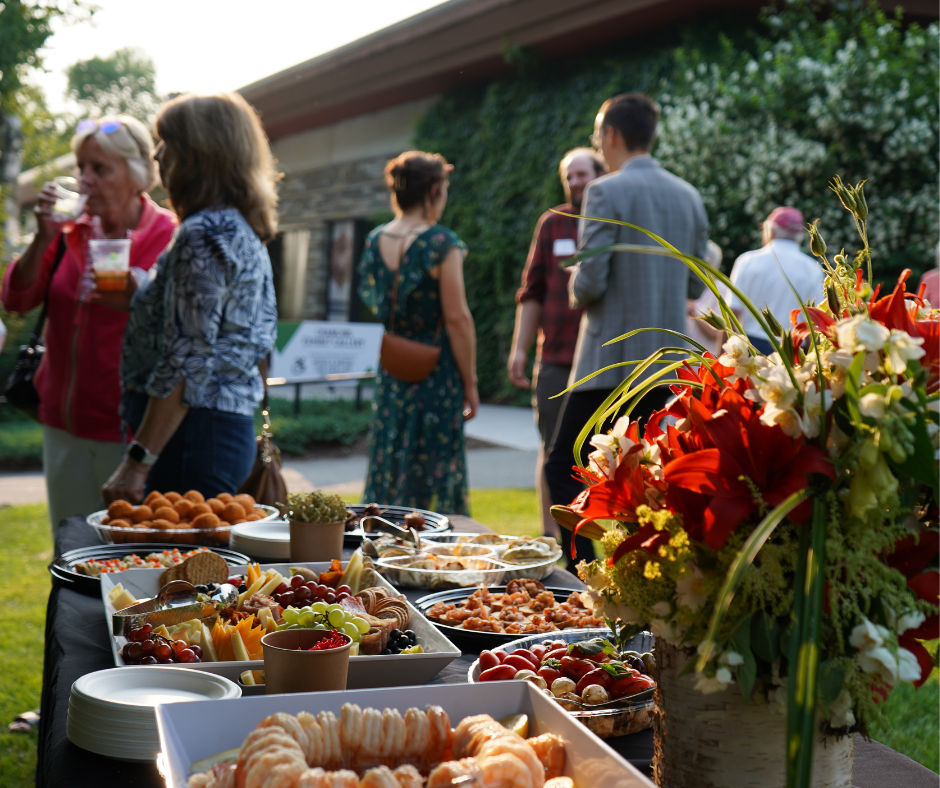 A group of people are standing around a table filled with food. The table is covered with a variety of dishes, including bowls, cups, and a vase. The people are engaged in conversation and enjoying the food.